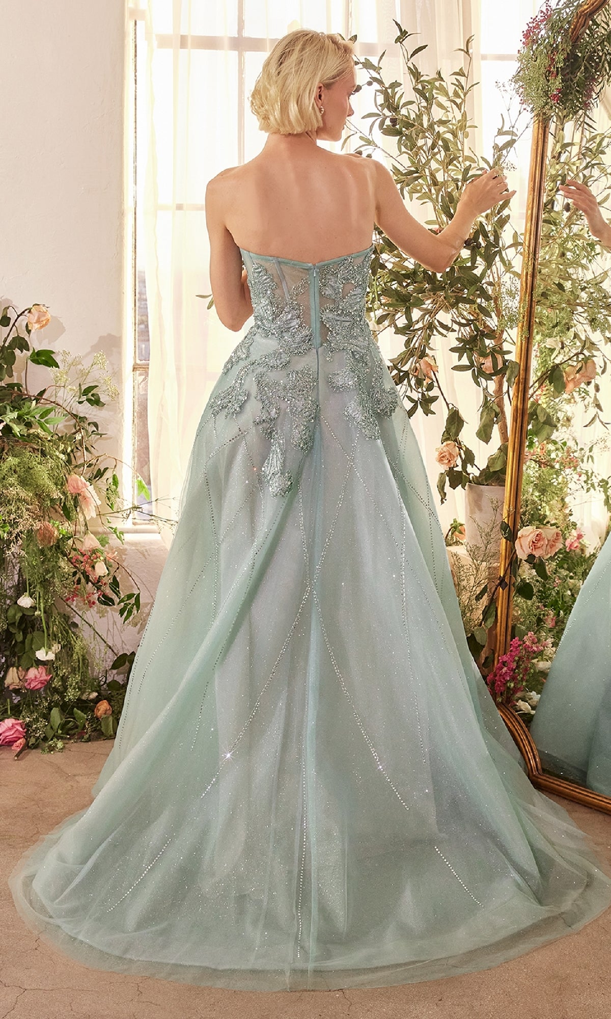 A woman wearing the Andrea & Leo A1387 Strapless Long Prom Ball Gown with lace detailing stands facing a mirror in a room decorated with greenery and flowers.